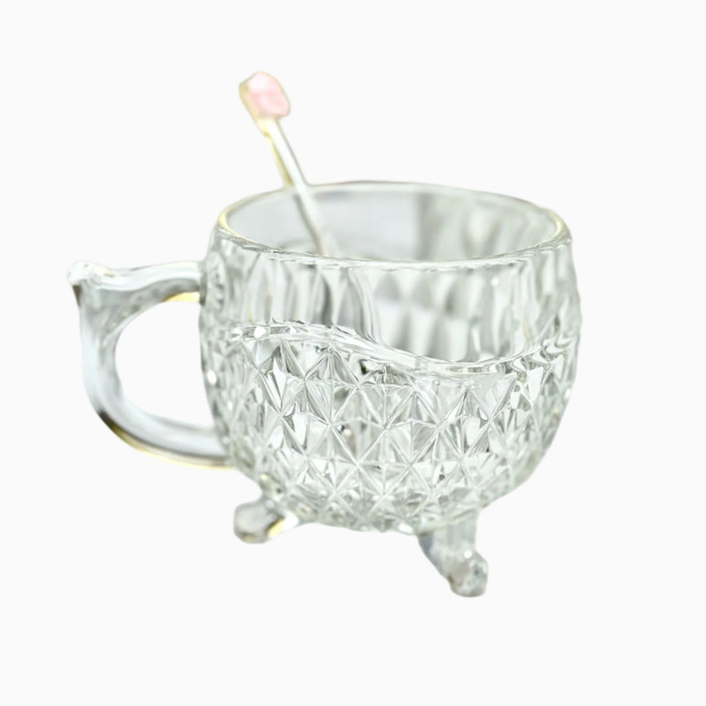 A clear glass tea cup with a geometric pattern and a handle, accompanied by a metal spoon with a pink tip.